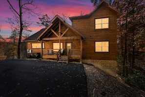 A wooden cabin with a porch, a driveway, and a forested backdrop. - New! 5BR w/Pool*Theater*Hot Tub*Fireplace*Games (Sevierville)