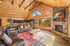 L’intérieur d’une cabane en rondins, avec un canapé en cuir, une cheminée et une grande fenêtre donnant sur une forêt.