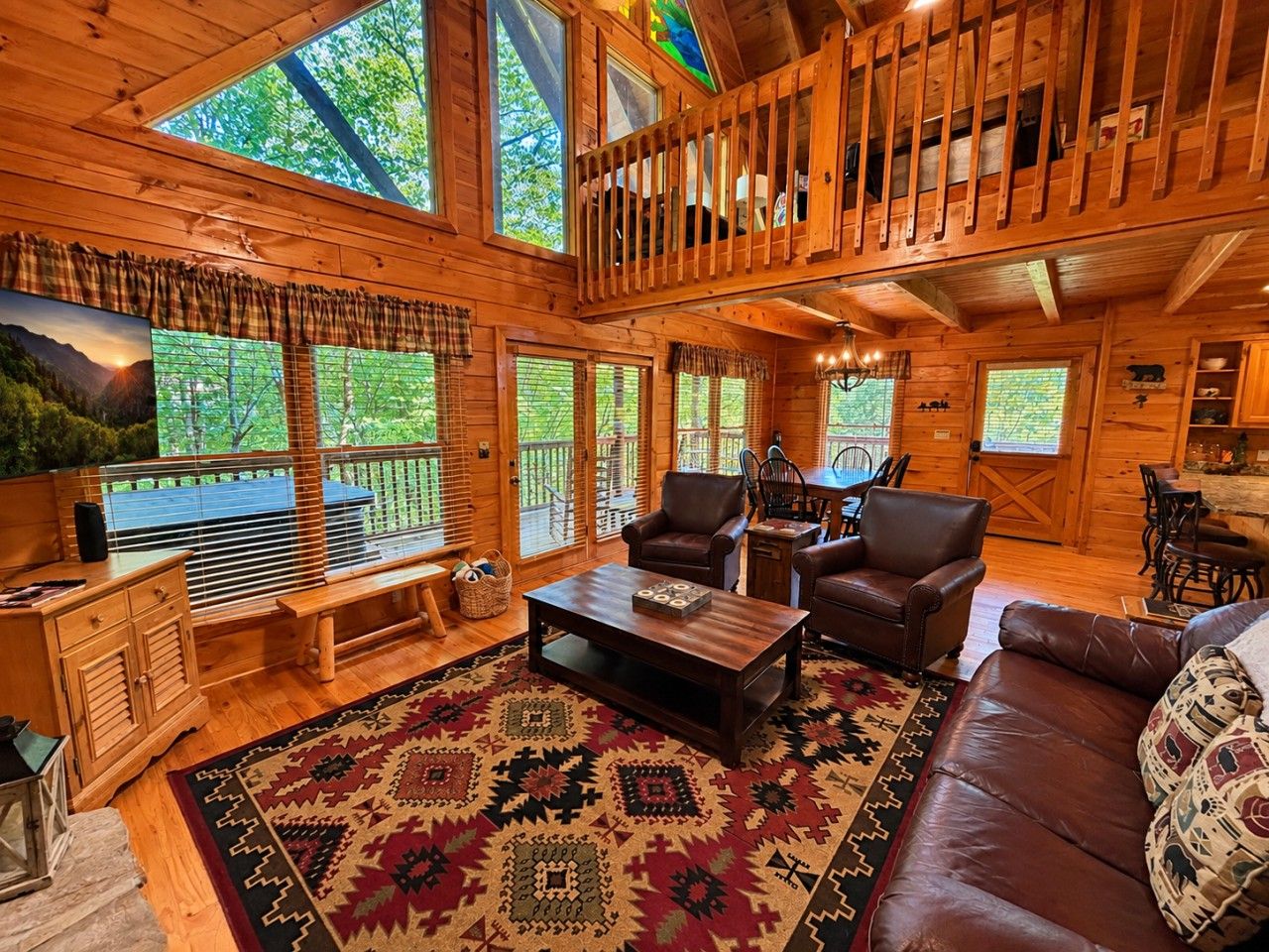 A cabin interior with a wooden floor, a large window overlooking a forest, a sofa, a coffee table, and a dining area.