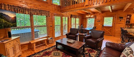 A cabin interior with a wooden floor, a large window overlooking a forest, a sofa, a coffee table, and a dining area.