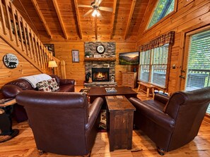 A cabin interior with a fireplace, leather furniture, wooden walls, and a view of the outdoors. - 2BR Resort Cabin w/Hot Tub*Game Room*Fireplaces (Pigeon Forge)