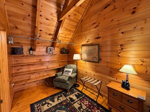 Ein gemütliches Blockhaus-Interieur mit Holzbalkendecke, einem gemusterten Sessel, einem kleinen Tisch mit Lampe und einem Teppich mit Tiermotiven.