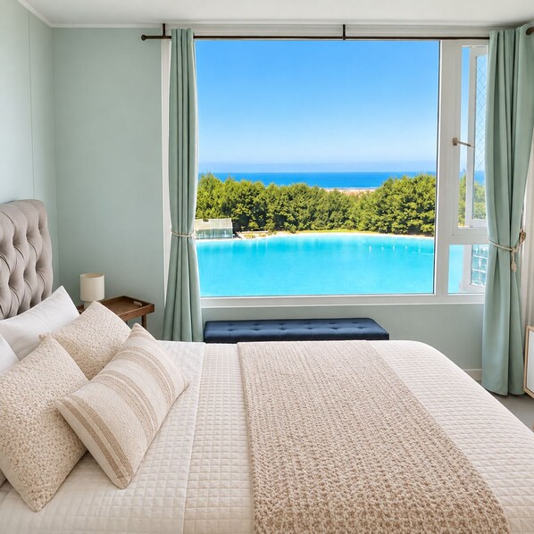 A bedroom with a large bed, a bedside table, and a view of a turquoise sea and green landscape.