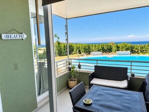 A balcony with a view of a swimming pool, a sign indicating 'BEACH', wicker furniture, and a potted plant.