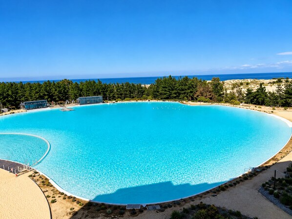 A large, clear blue swimming pool with a curved edge, surrounded by a sandy beach and dense greenery.