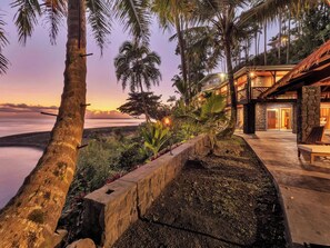 A beachfront property with a stone retaining wall, palm trees, and a building with a thatched roof. - Honolii Hale - Oceanfront (Hilo)