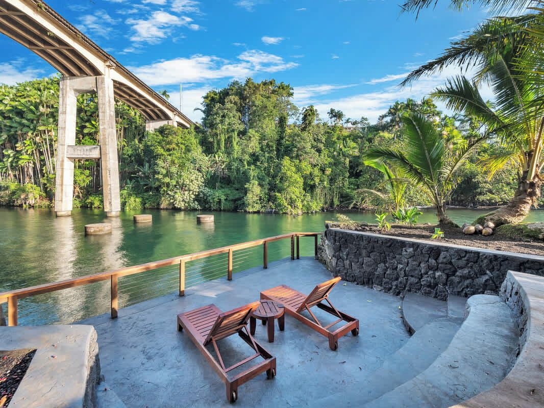 A concrete deck with lounge chairs and a table overlooking a river, with a bridge in the background.