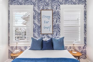 A bedroom with a bed, two bedside tables, a framed sign, and a window with blinds. - The Good Life (Santa Rosa Beach)