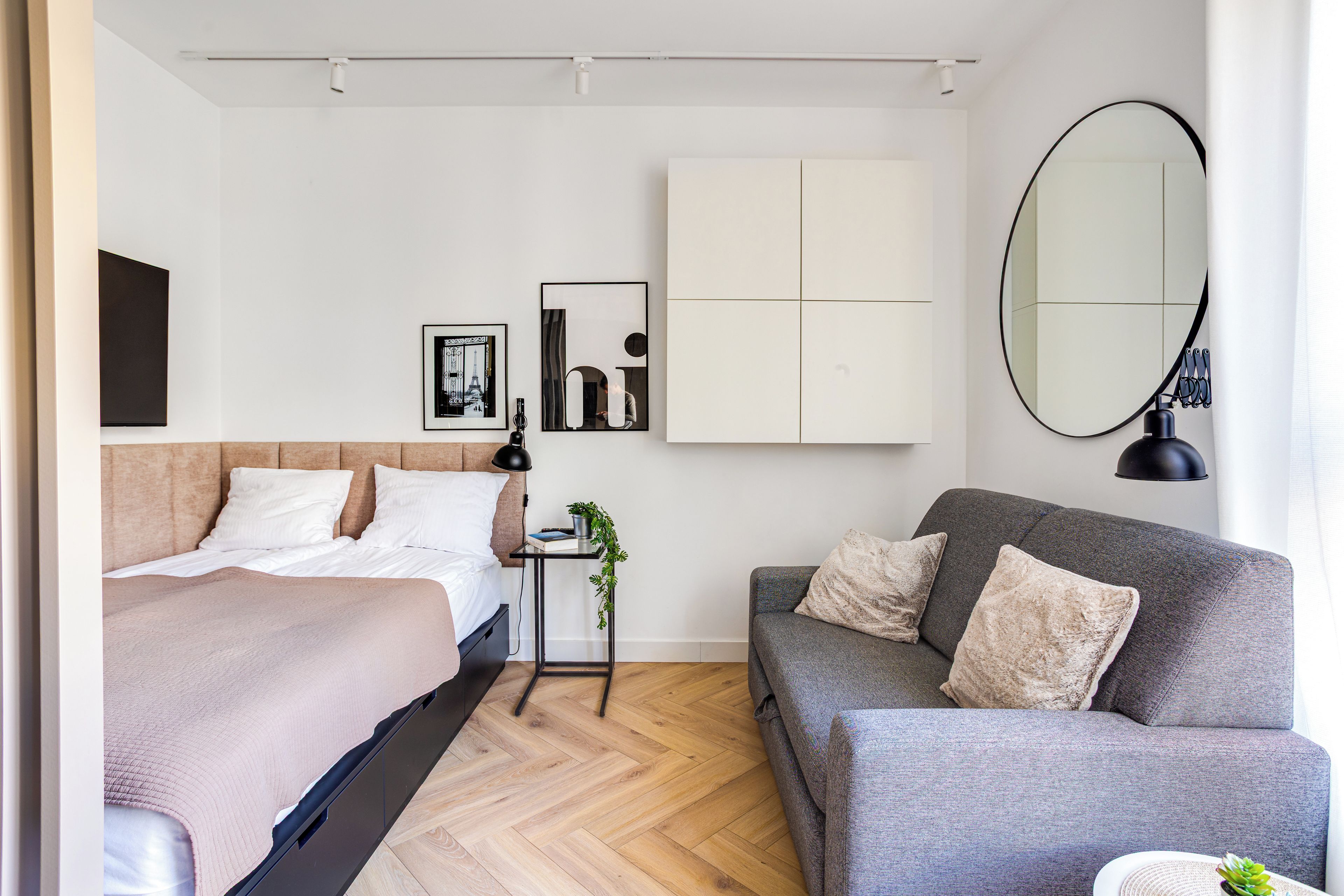 Un dormitorio moderno con cama, sofá gris, una mesita de noche con planta y un espejo grande en la pared.