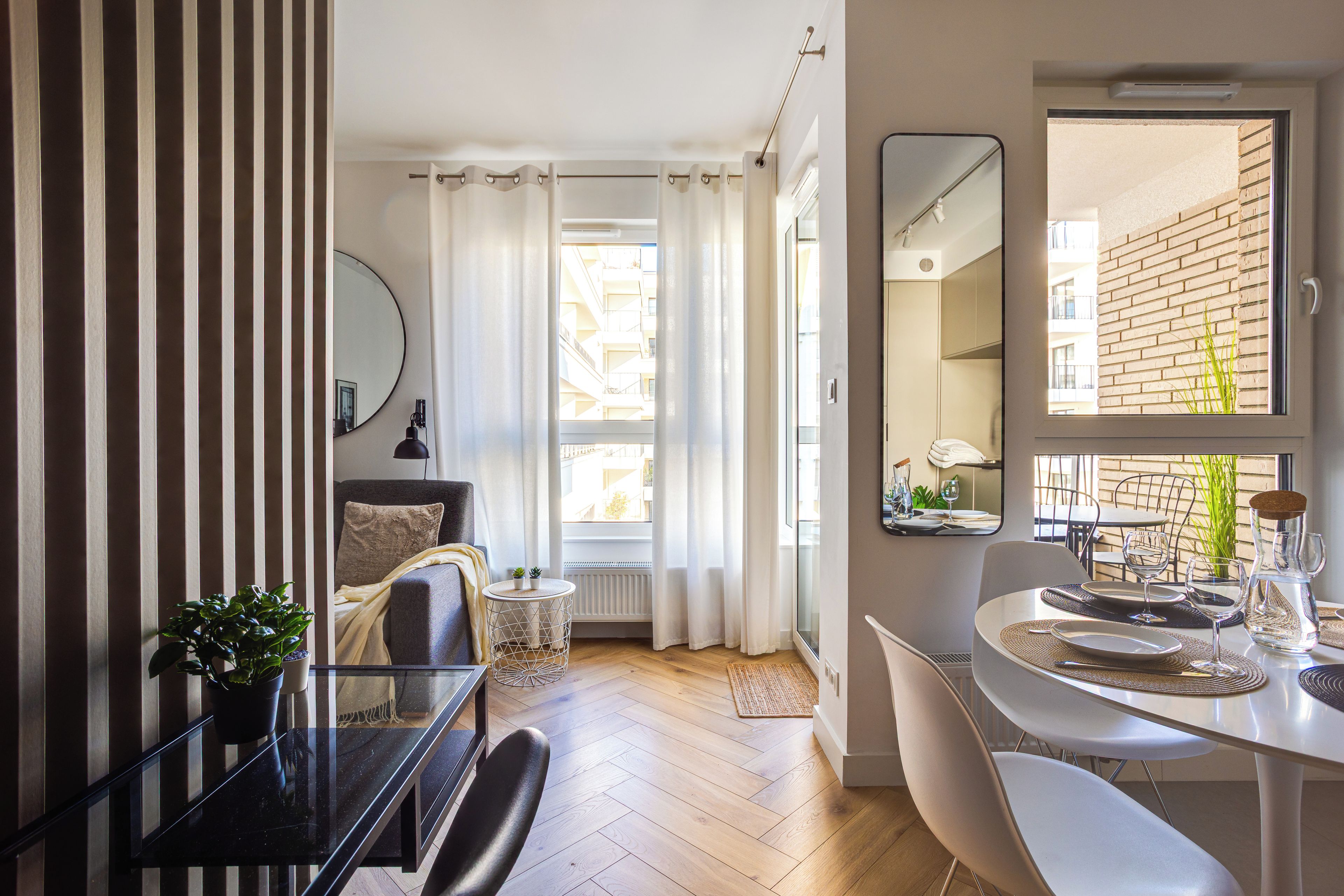 A modern living room with a dining area, a large window with curtains, a potted plant, and a mirror on the wall.