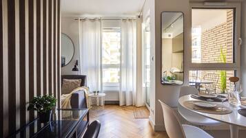 A modern living room with a dining area, a large window with curtains, a potted plant, and a mirror on the wall.