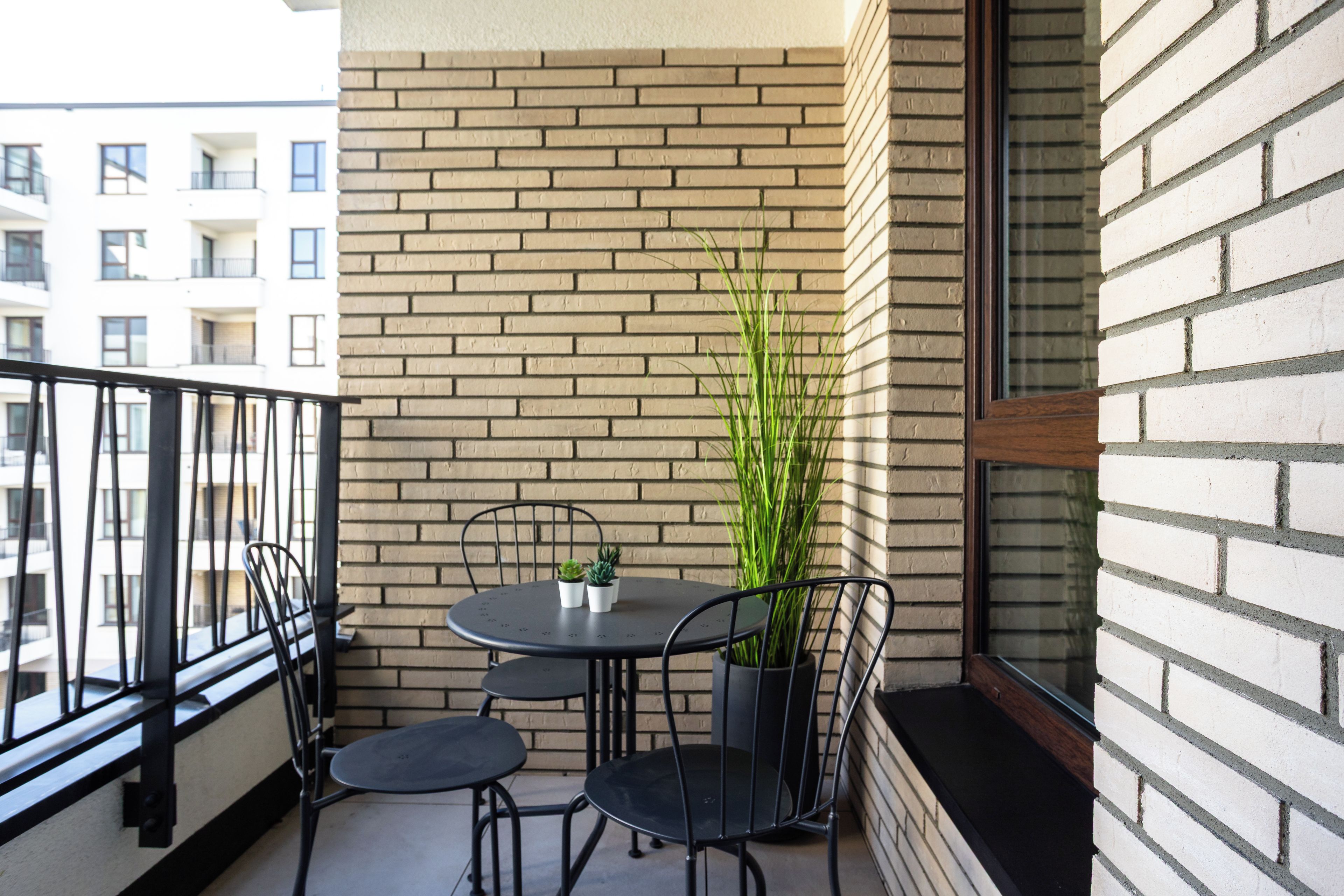 Een balkon met een bakstenen muur, een ronde tafel met stoelen, een bloempot en een raam.