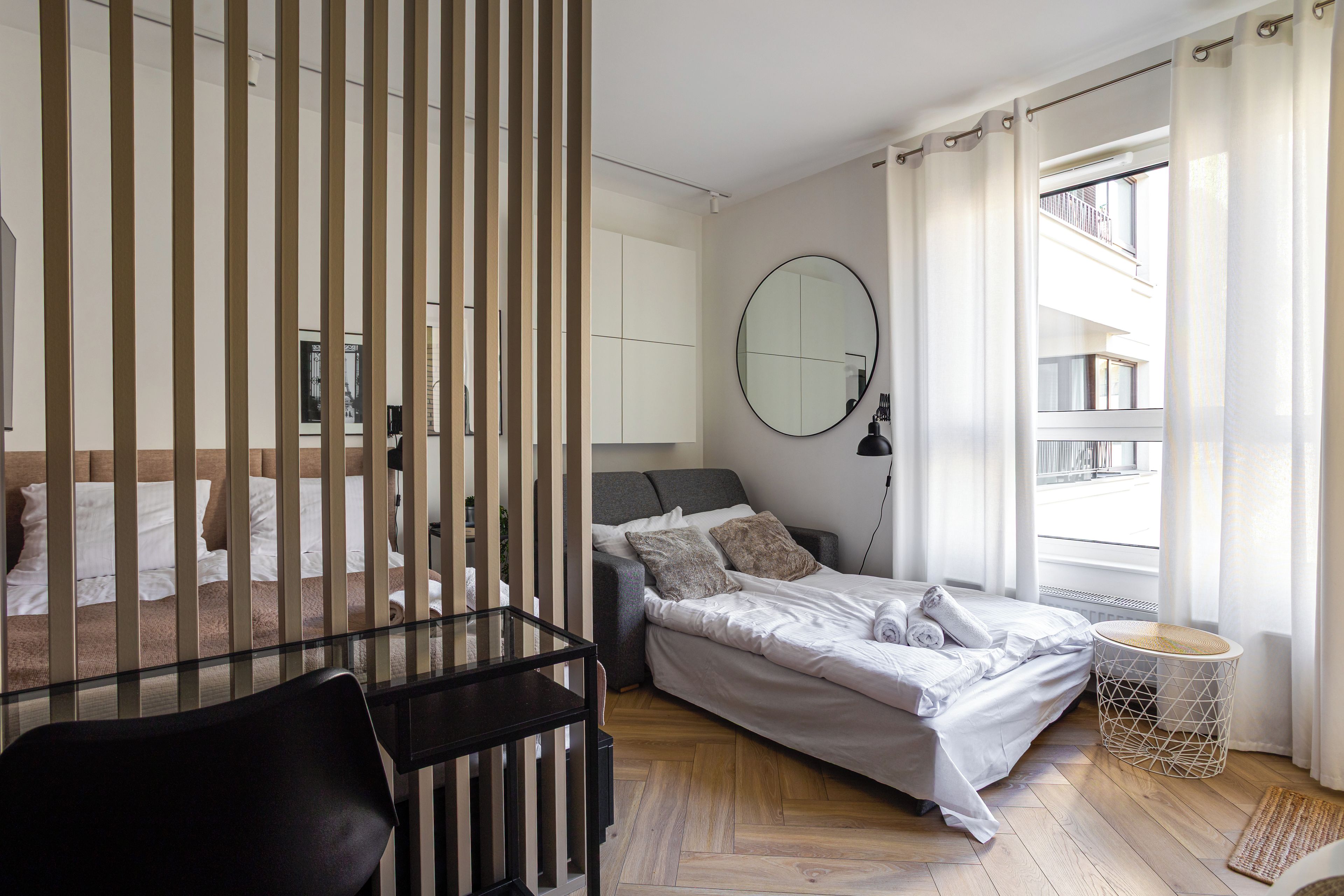 A modern bedroom with a bed, a desk, a chair, a round mirror, and a window with curtains.