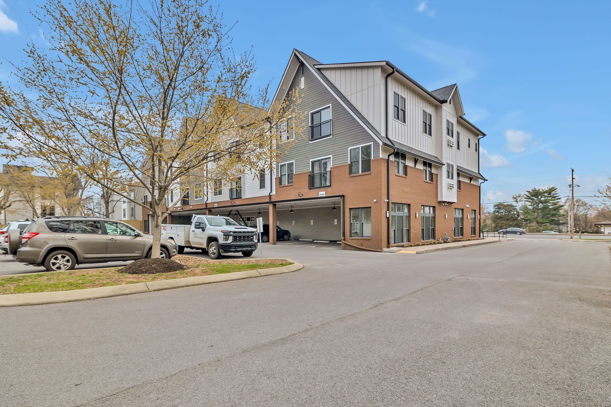 A multi-story residential building with a garage, parked cars, and a tree in the driveway.