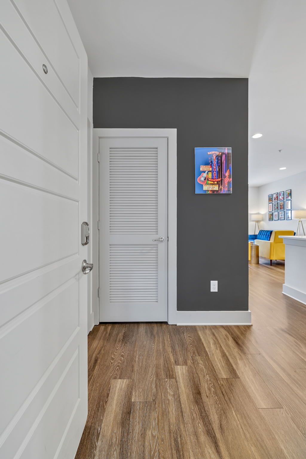 A hallway with a white door, grey walls, and wooden flooring. There is a colorful poster on the wall and a yellow chair in the background.