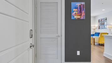 A hallway with a white door, grey walls, and wooden flooring. There is a colorful poster on the wall and a yellow chair in the background.