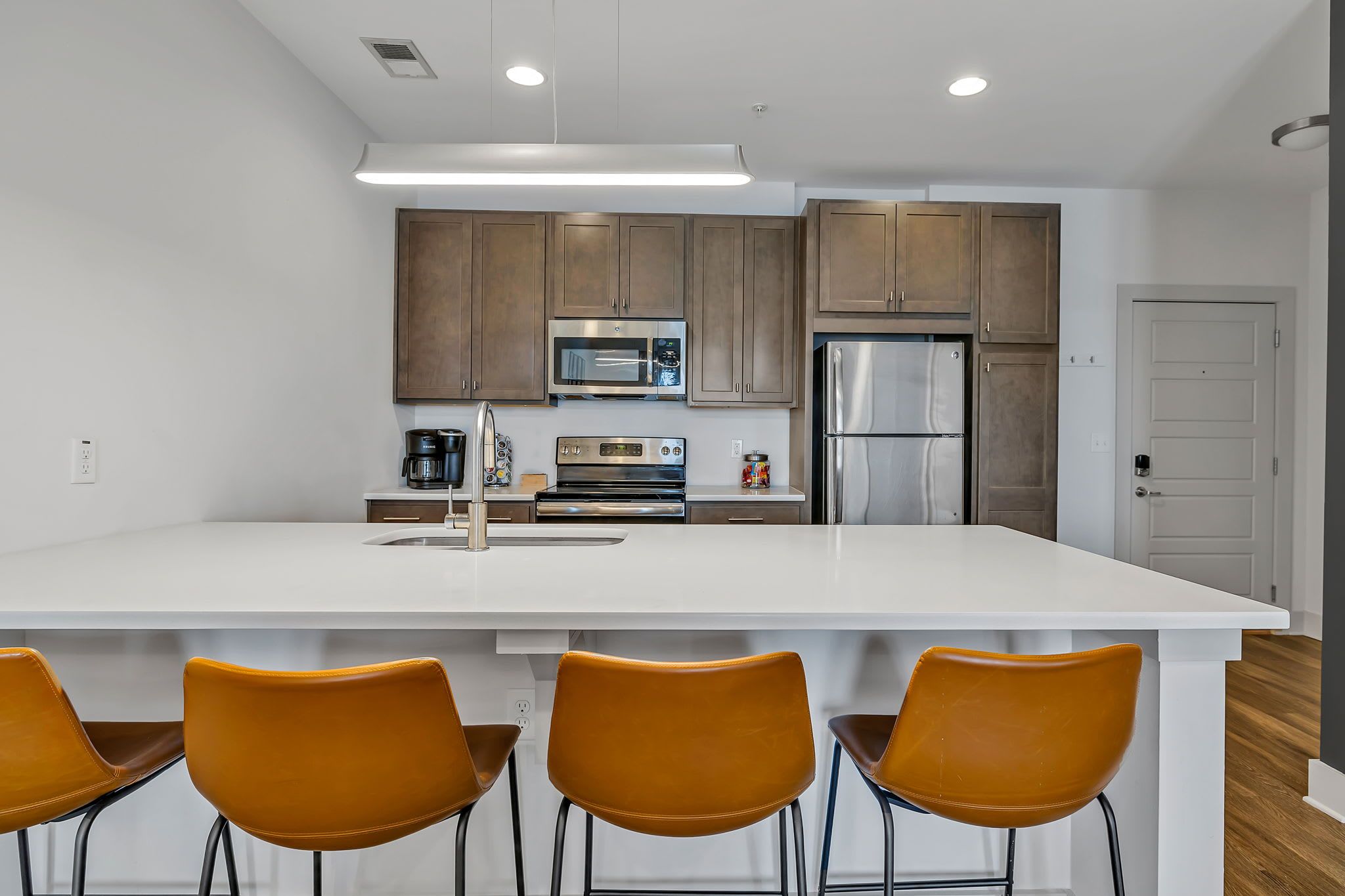 A modern kitchen with white countertops, brown cabinets, stainless steel appliances, and three orange chairs.