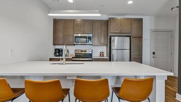 A modern kitchen with white countertops, brown cabinets, stainless steel appliances, and three orange chairs.