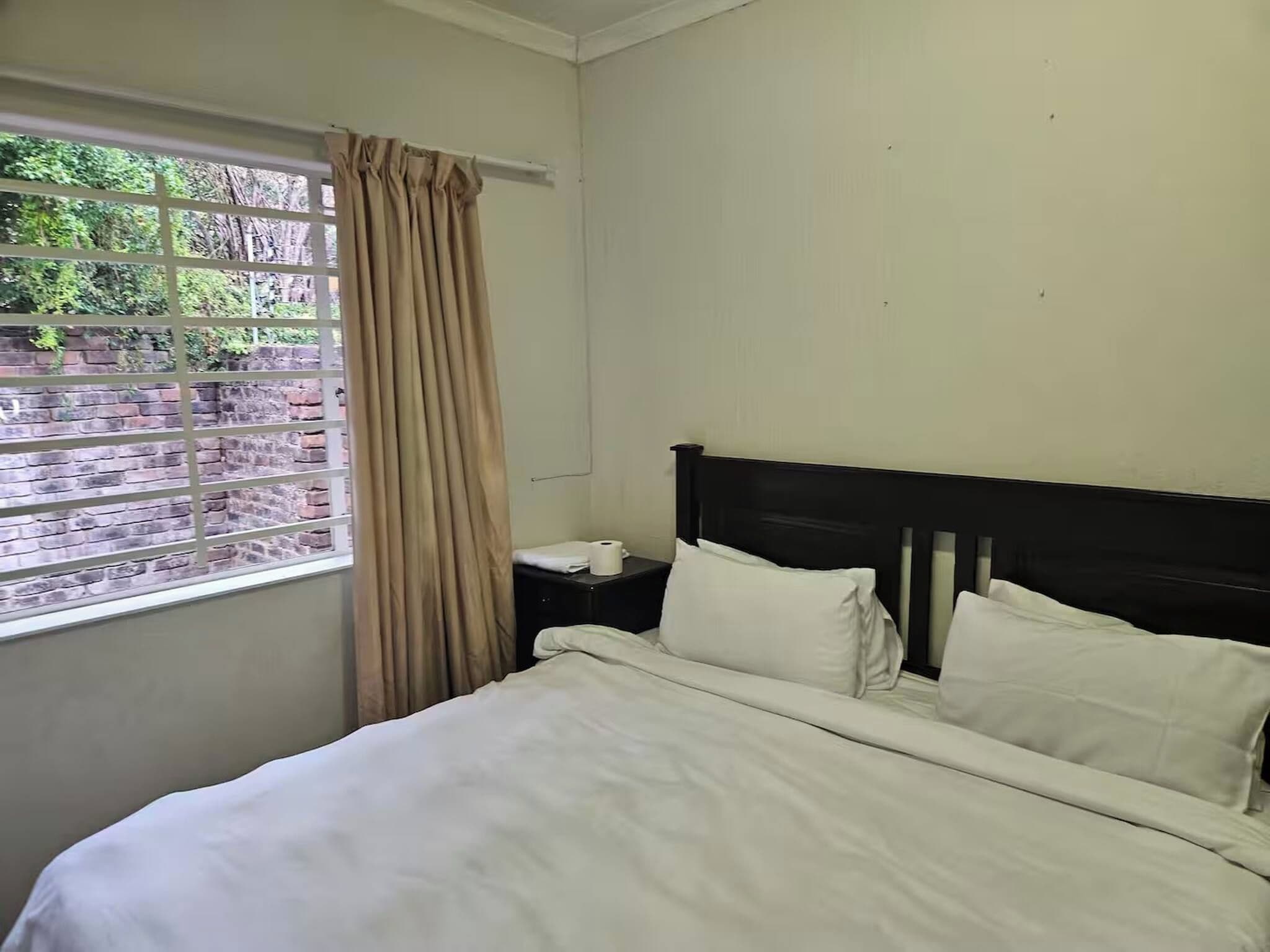 A hotel room with a bed, a nightstand, a window with curtains, and a view of greenery outside.