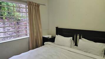 A hotel room with a bed, a nightstand, a window with curtains, and a view of greenery outside.