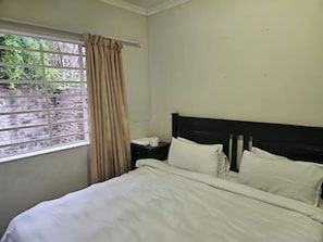 A hotel room with a bed, a nightstand, a window with curtains, and a view of greenery outside. - Spacious 3-bedroom House Rental in Randburg, South Africa (Johannesburg)