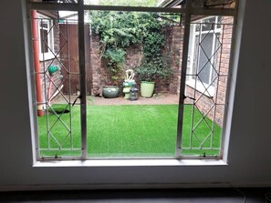 A view through a window with a metal gate leading to a backyard with a brick wall, potted plants, and a statue. - Spacious 3-bedroom House Rental in Randburg, South Africa (Johannesburg)