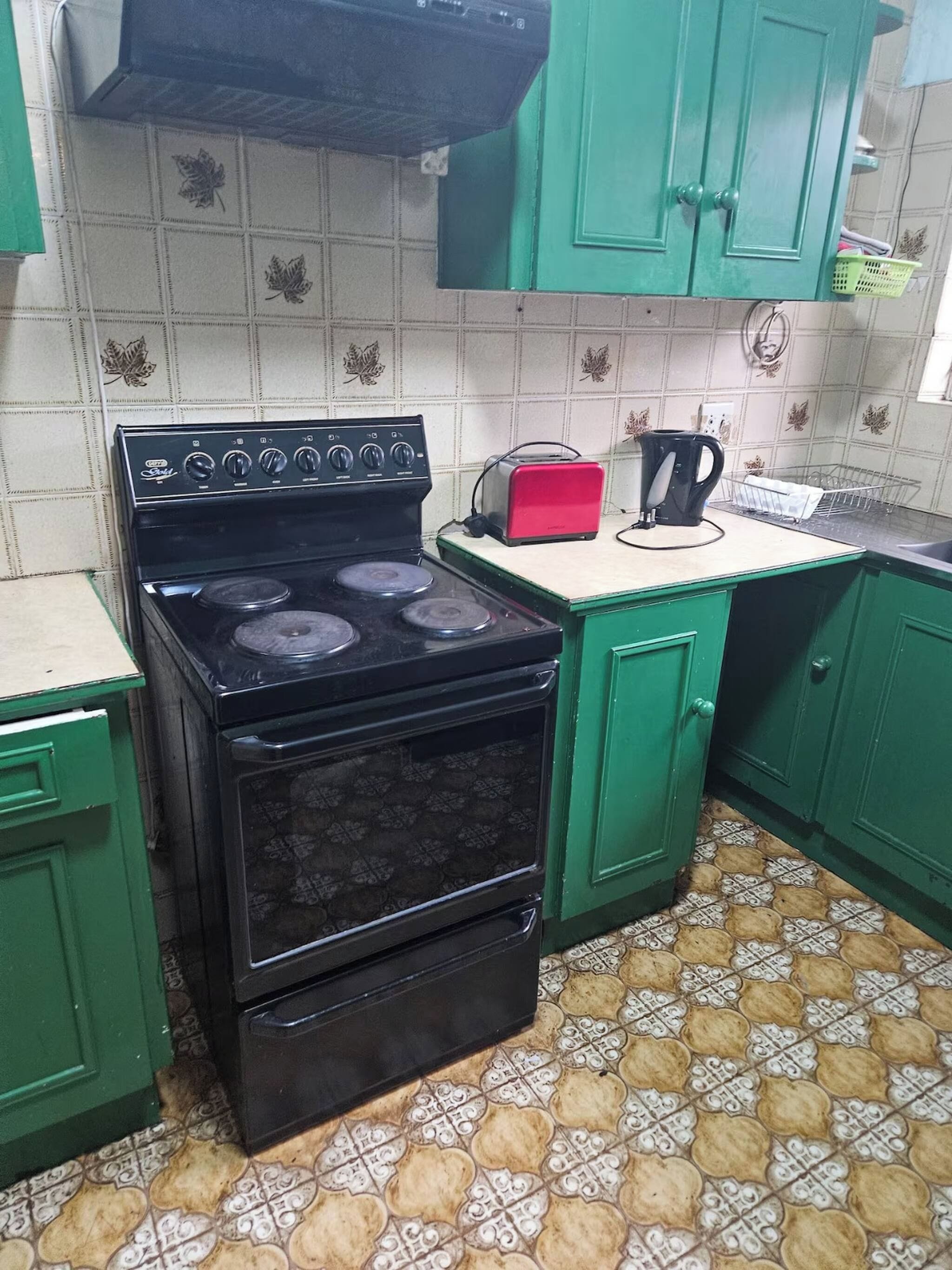A kitchen with a black stove, green cabinets, a red toaster, and patterned floor tiles.