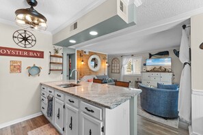 A kitchen with a central island, a dining area with a blue chair, and a wall-mounted TV. - Porch & Palms - ground-floor 2BR, pool, near beach (North Myrtle Beach)