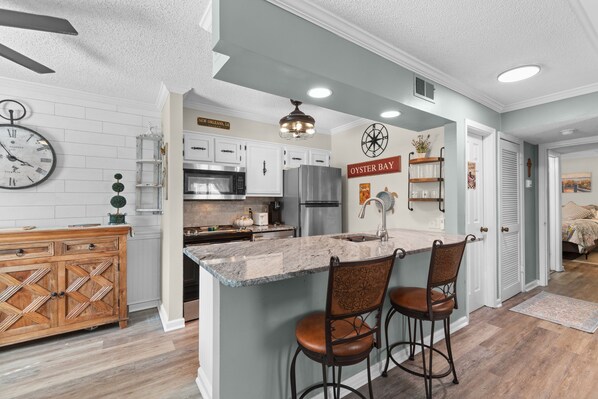 A kitchen with a central island, bar stools, a clock, and a sign reading 'Oyster Bay.' - Porch & Palms - ground-floor 2BR, pool, near beach (North Myrtle Beach)