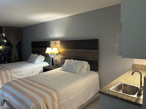 Habitación de hotel con dos camas, una mesita de noche con lámpara, una kitchenette con lavamanos y vistas al balcón.