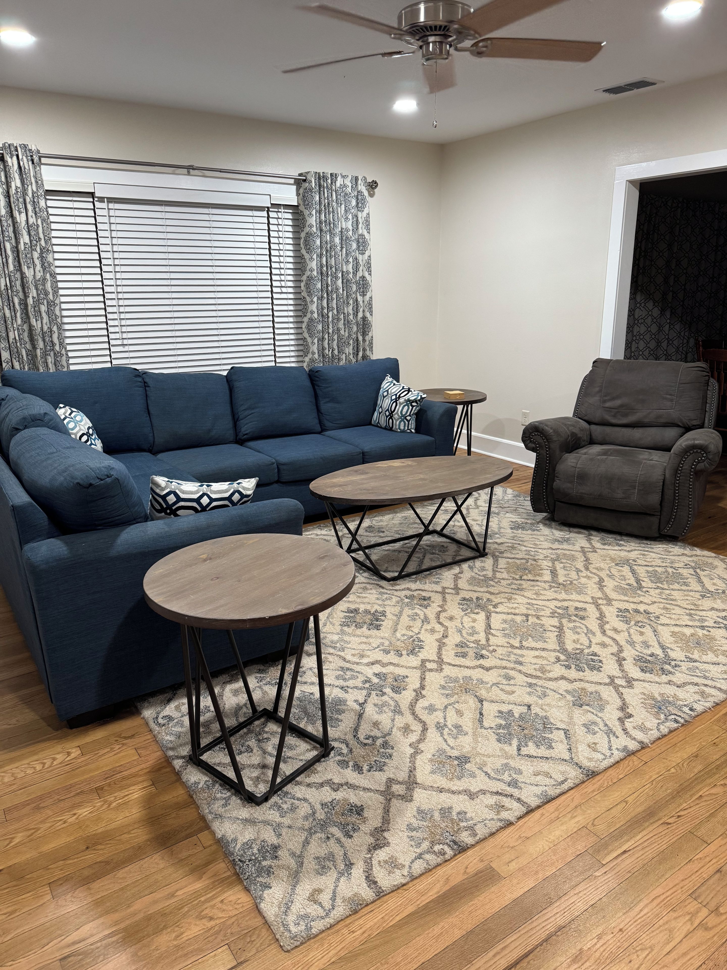 Ein Wohnzimmer mit einem blauen Sofa, zwei runden Couchtischen, einem gemusterten Teppich und einem Deckenventilator.