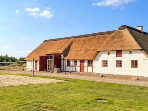 Ein traditionelles reetgedecktes Haus mit weißer Fassade und roten Akzenten, eingebettet in eine grüne Umgebung unter blauem Himmel.