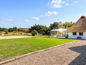 Ein Haus mit strohgedecktem Dach und einem Wintergarten, umgeben von einer Schotterauffahrt und einer Rasenfläche.