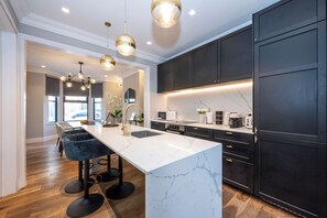 Sleek, modern kitchen featuring dark cabinetry, a striking white marble waterfall island, elegant gold pendant lighting, and plush blue velvet barstool seating. - Family Home by Battersea Park | 4 Beds + Garden (Greater London)
