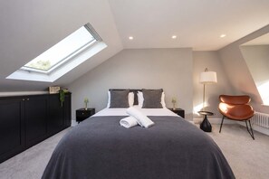 A modern bedroom with a skylight, a large bed, a chair, and a nightstand. - Family Home by Battersea Park | 4 Beds + Garden (Greater London)