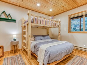 A bunk bed with a wooden frame, a bedside table with a lamp, and a window with a view of trees.