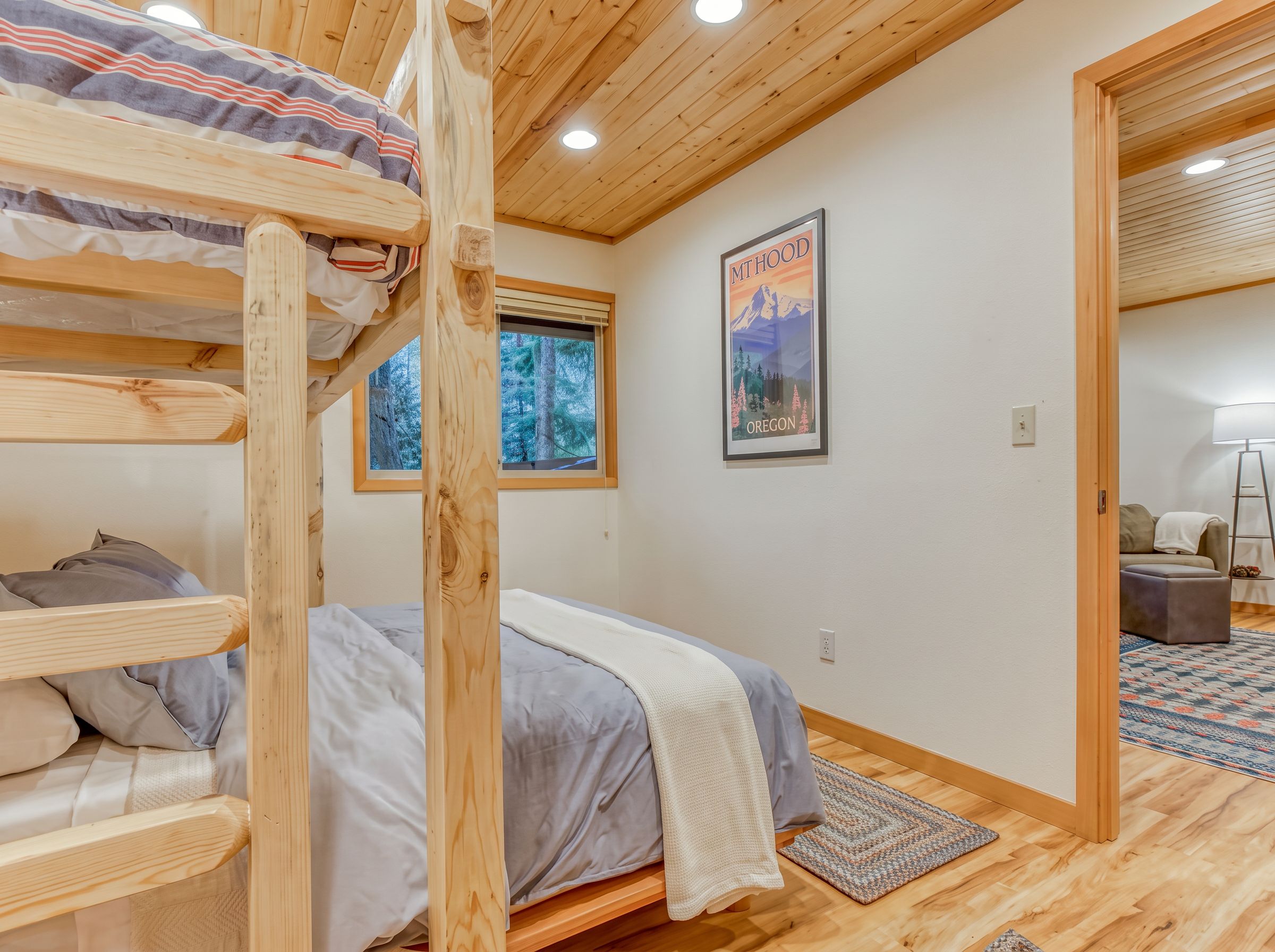 A room with a bunk bed, a framed poster, a window with a view of trees, and a wooden floor.