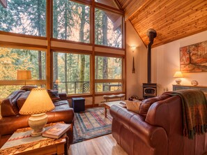 A cozy living room with leather sofas, a fireplace, and large windows offering a forest view. - Sandy Riverfront Retreat • Dogs OK • Ski & Hike (Rhododendron)