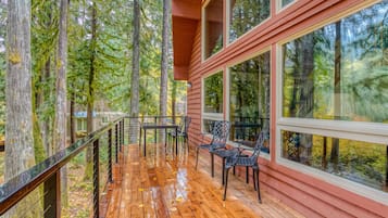A wooden deck with a railing, a table, and chairs, overlooking a forested area.