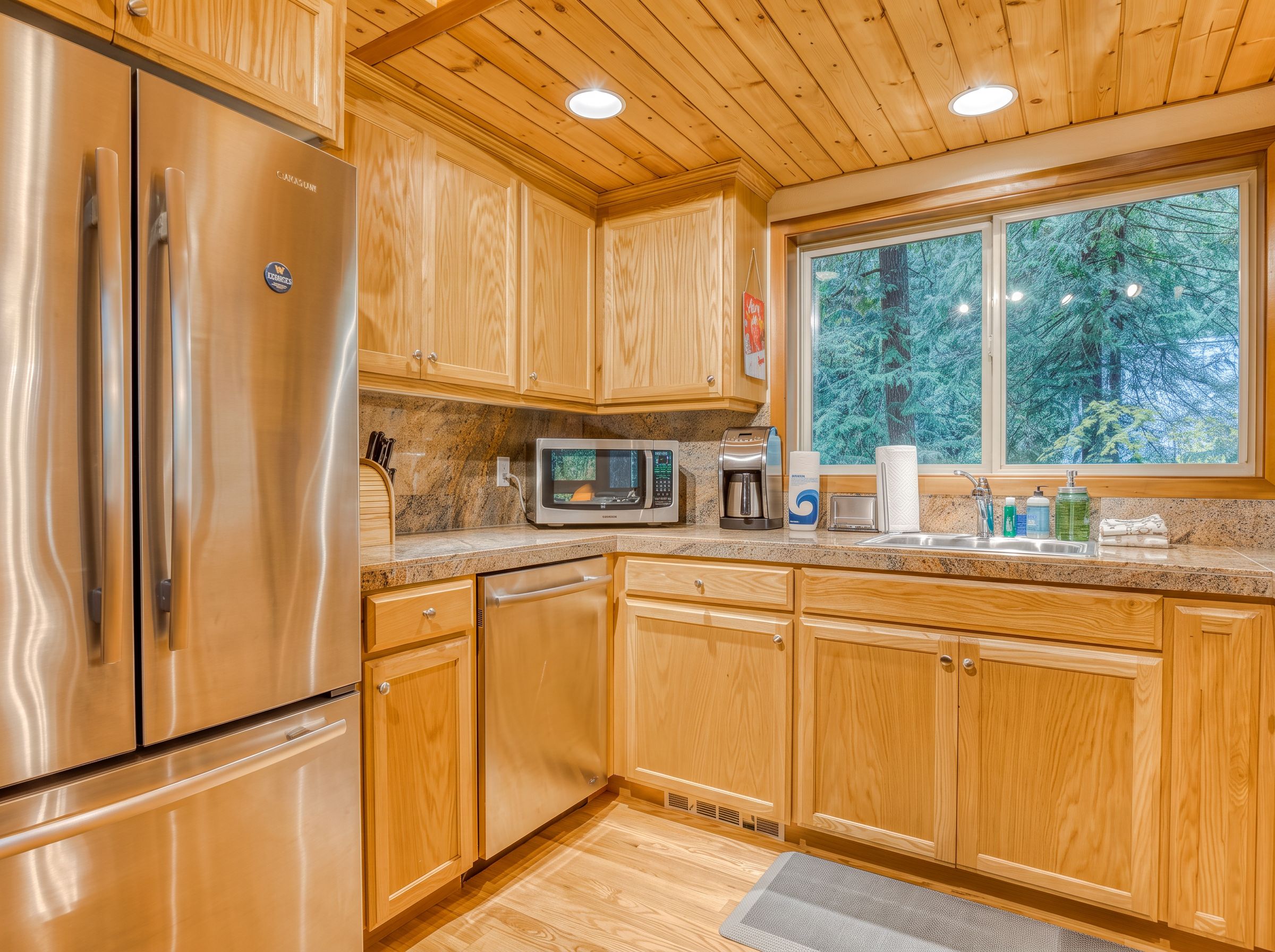 A kitchen with wooden cabinets, stainless steel appliances, and a window with a forest view.