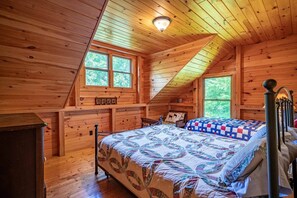 A wooden cabin bedroom with a sloped ceiling, two beds with quilts, a fireplace, and a window overlooking greenery. - Camp Shanahan - Boat Access only camp (Webb)