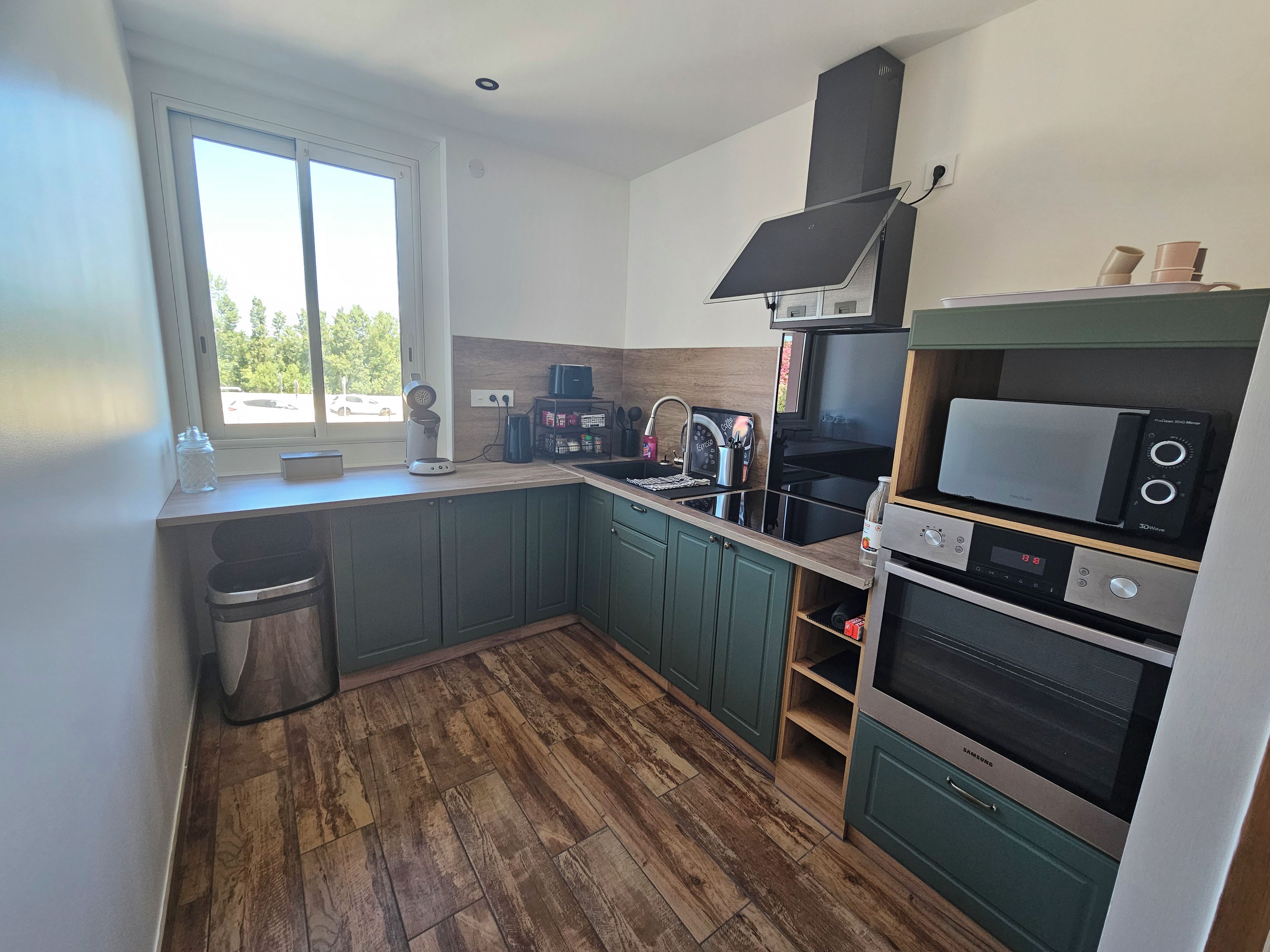 A kitchen with a window, wooden flooring, green cabinets, a sink, a stove, and a microwave.