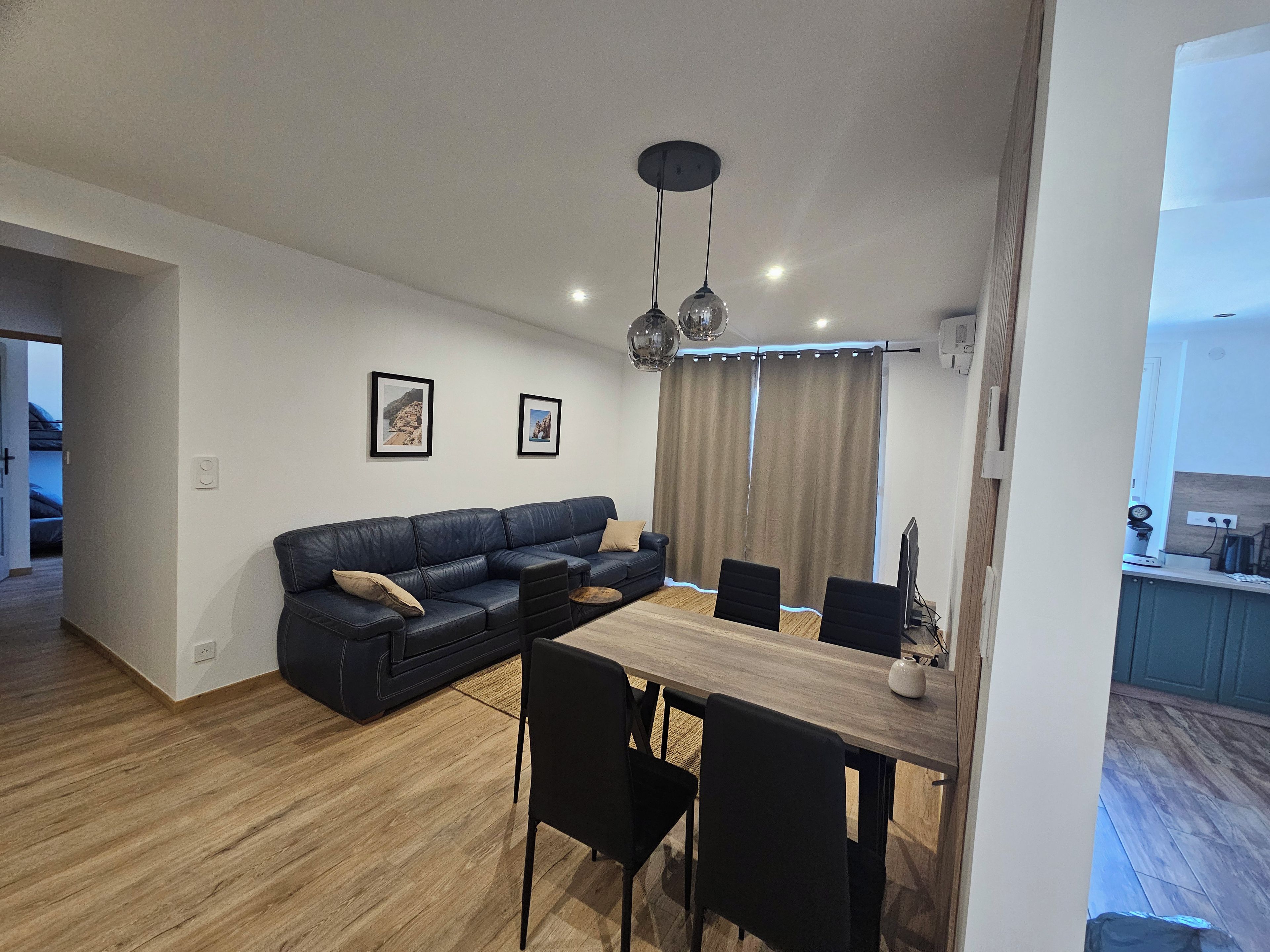 A modern living space with a sectional sofa, dining table, and chairs. There are two framed pictures on the wall, a ceiling light fixture, and a window with curtains.