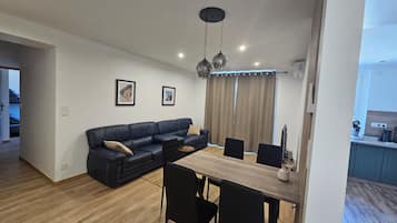 A modern living space with a sectional sofa, dining table, and chairs. There are two framed pictures on the wall, a ceiling light fixture, and a window with curtains.