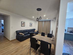 A modern living space with a sectional sofa, dining table, and chairs. There are two framed pictures on the wall, a ceiling light fixture, and a window with curtains. - Comfortable apartment 5 minutes from the train station (Perpignan)