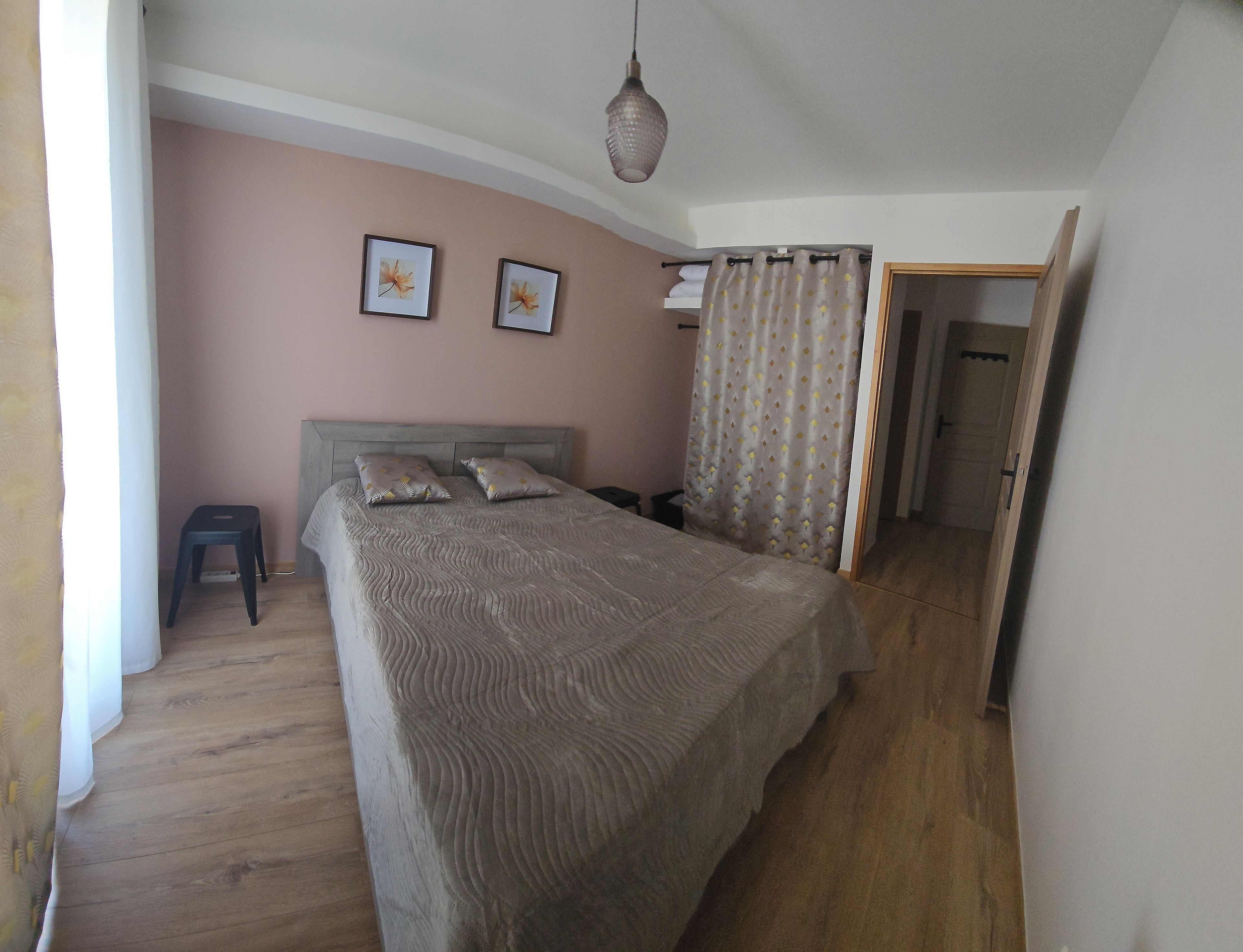 Quarto com cama, duas almofadas, cabeceira, mesa de cabeceira, luminária suspensa, porta e cortina.