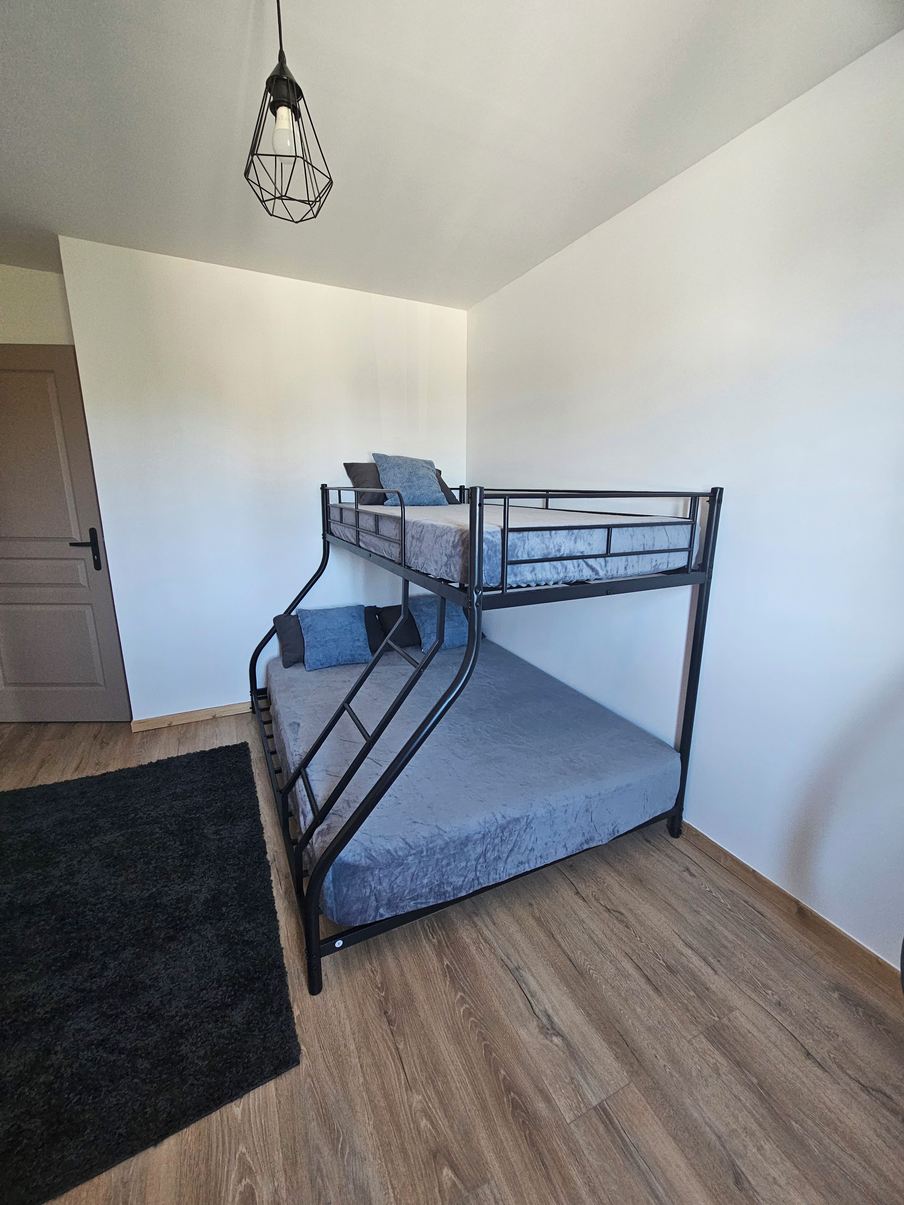 A bunk bed with a metal frame, a grey mattress, and a black headboard. There is a black rug on the floor and a door to the left.