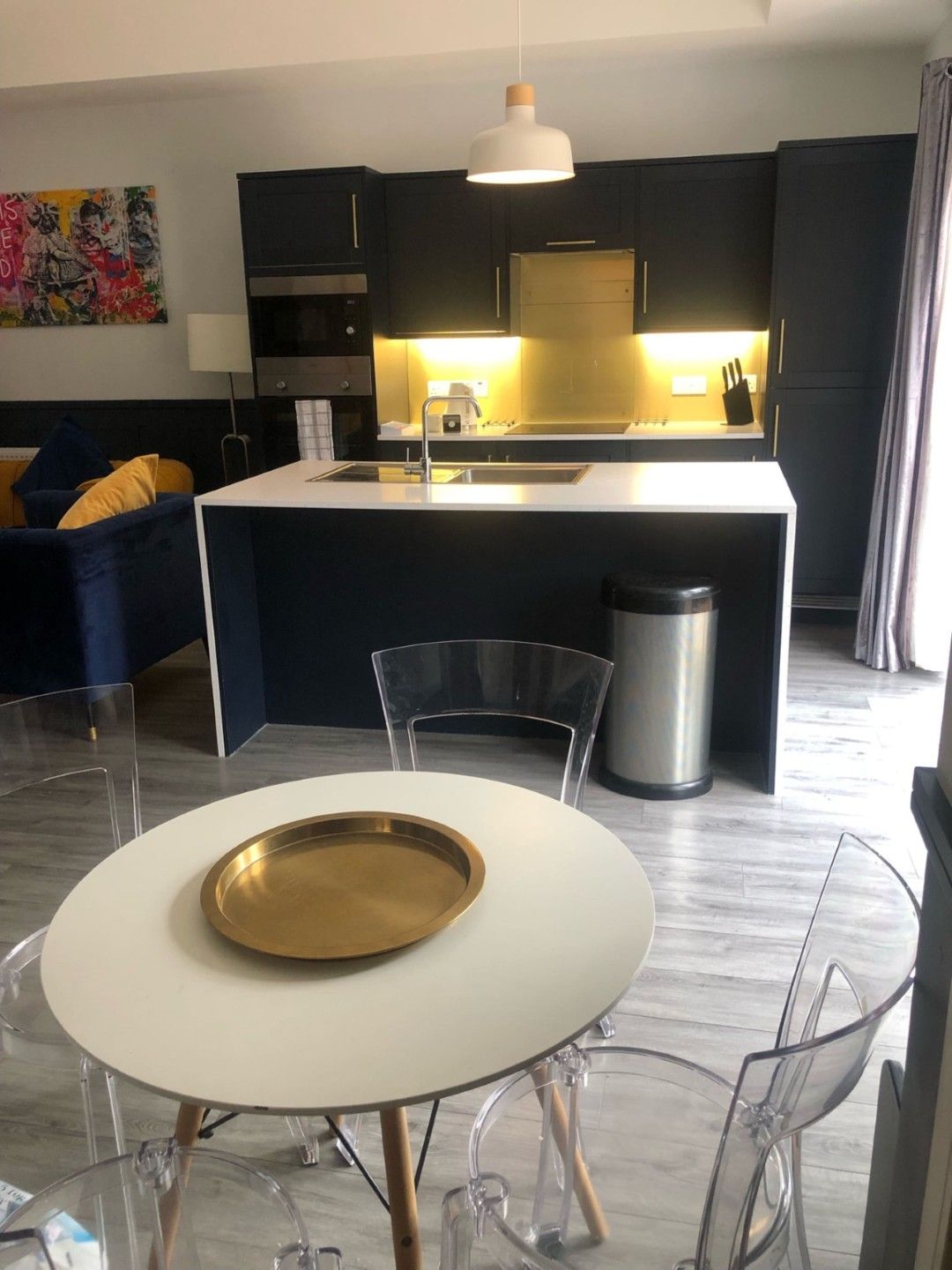A modern kitchen with a central island, a dining area with a white table and chairs, and a colorful abstract painting on the wall.