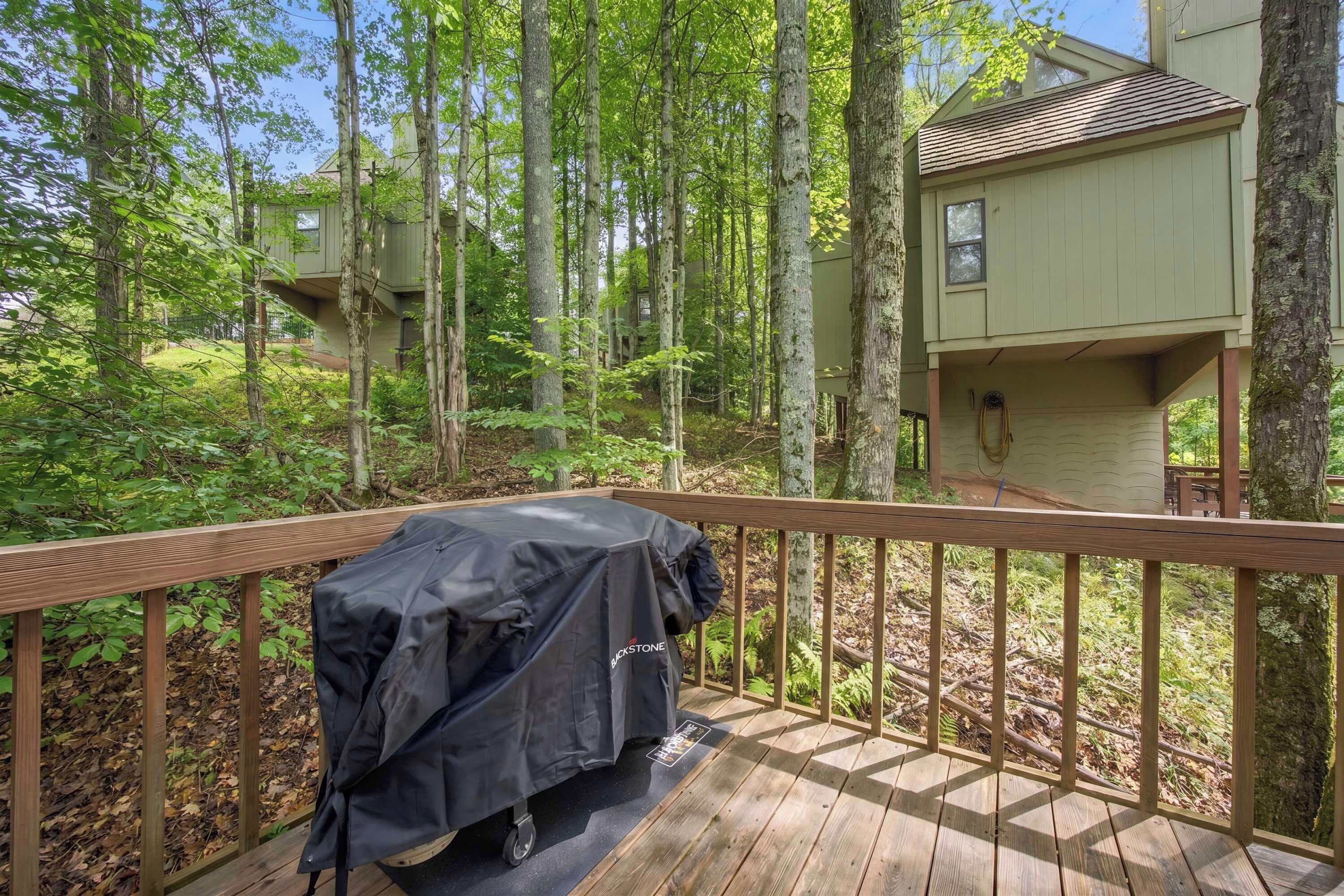 A wooden deck with a barbecue grill, surrounded by trees and a house in the background.