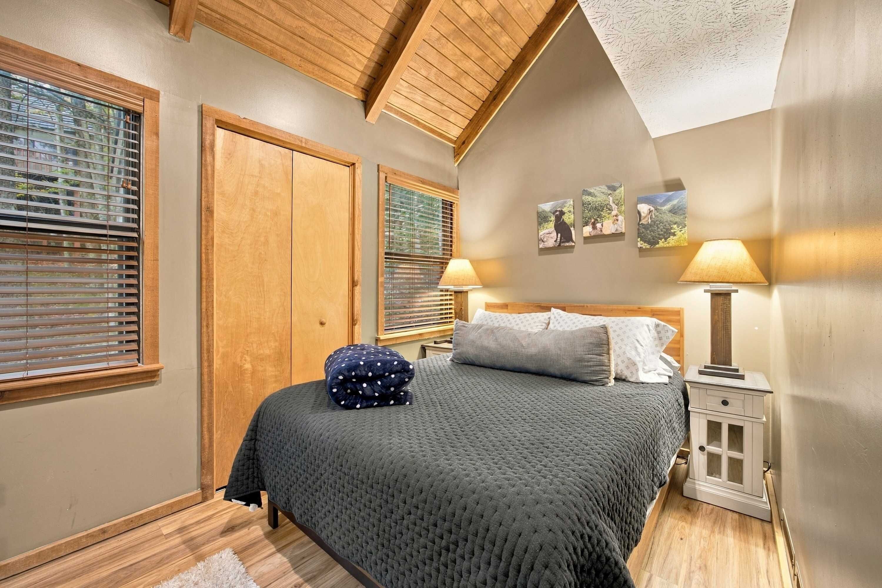 A bedroom with a bed, two windows with blinds, a nightstand, a lamp, and wall art.